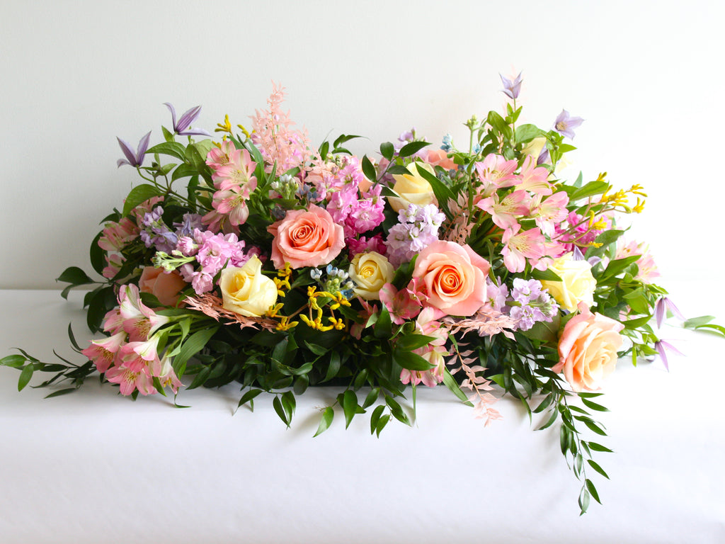 A massed casket spray of soft pastel flowers in peach, pink, soft yellow and lavender is set on a white background.