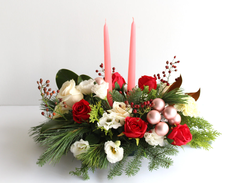 A table centerpiece is set against a white background. It is filled with red and white flowers and winter greenery and has two candles in the centre.