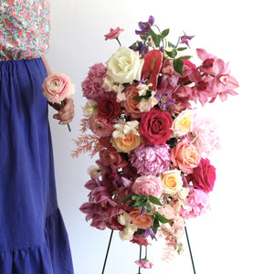 A funeral flower spray made with massed flowers in shades of pinks and creams, with a woman standing beside holding a single flower.