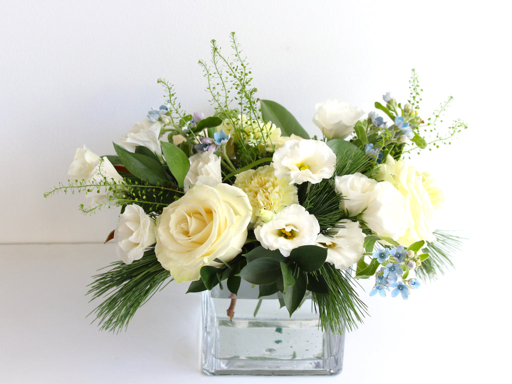 A glass vase filled with white and off white flowers with some pale blue blooms and winter greenery.