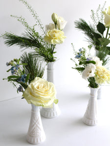 A set of four white budvases each has a cluster of white and blue flowers with greenery.