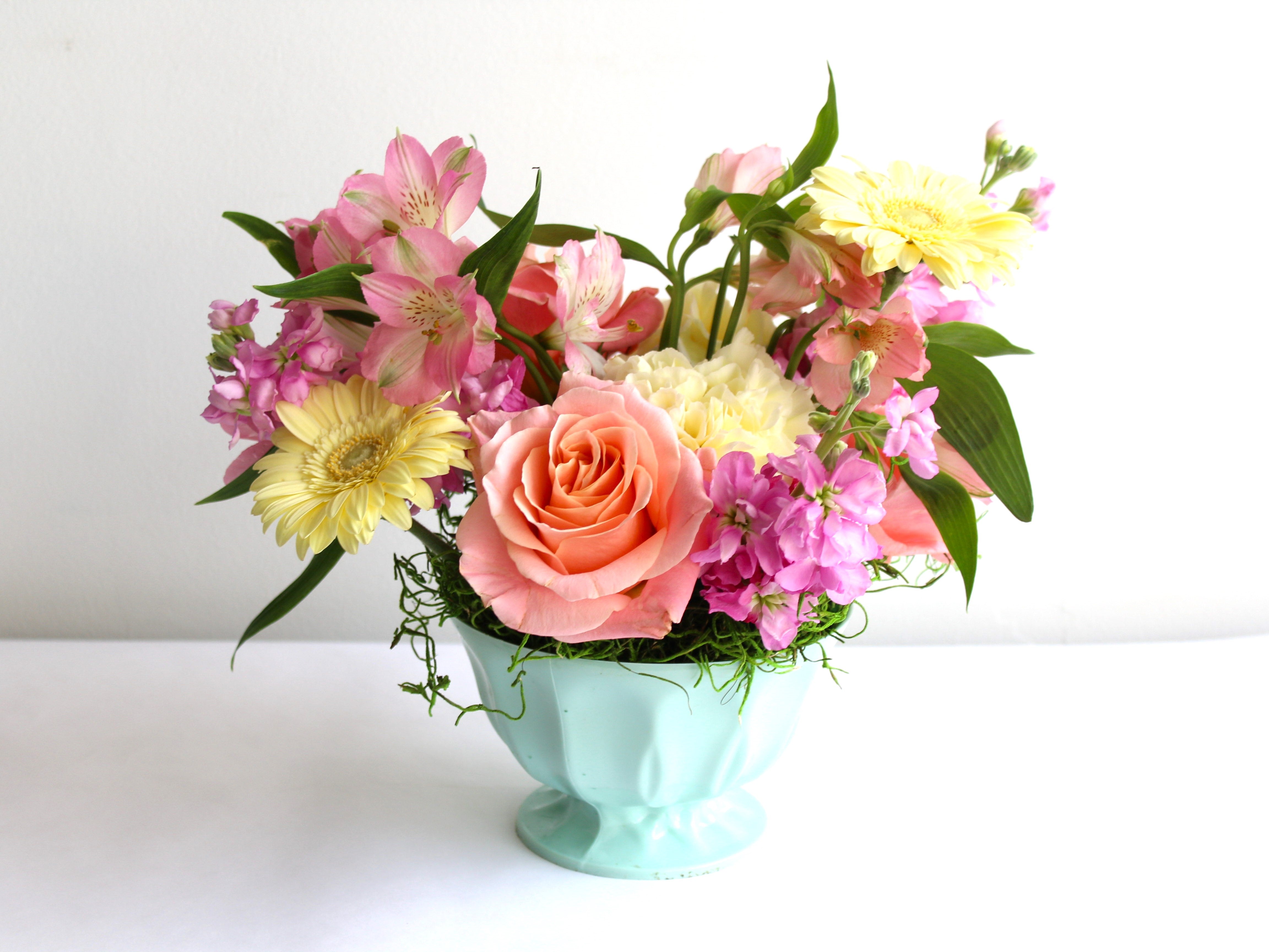 A mint coloured small dish has an arrangement of flowers in soft yellows, peach and pink.