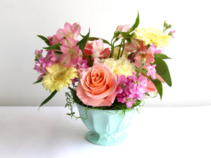 A mint coloured small dish has an arrangement of flowers in soft yellows, peach and pink.