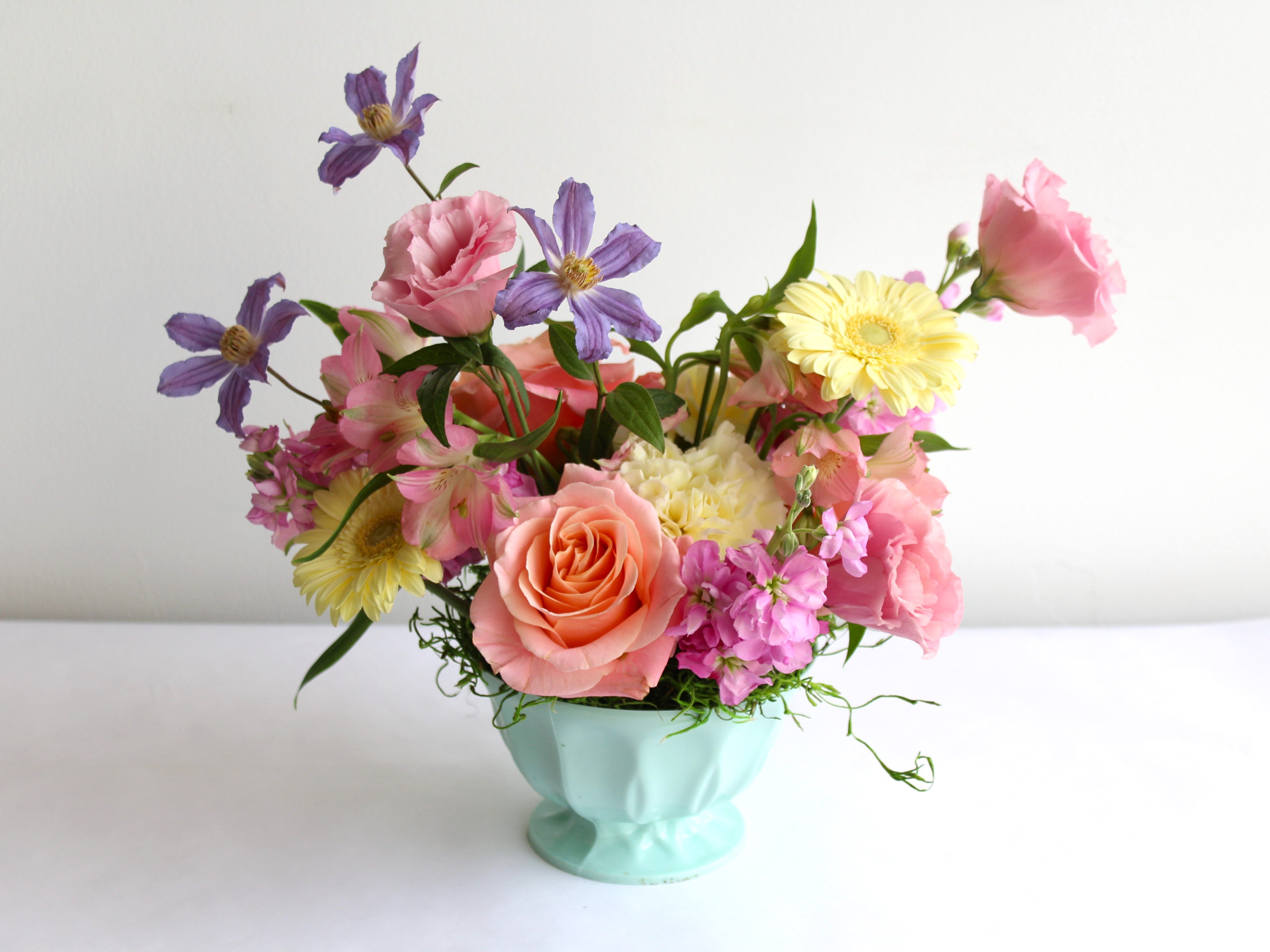 A mint coloured footed dish is holding an arrangement of pastel toned flowers.