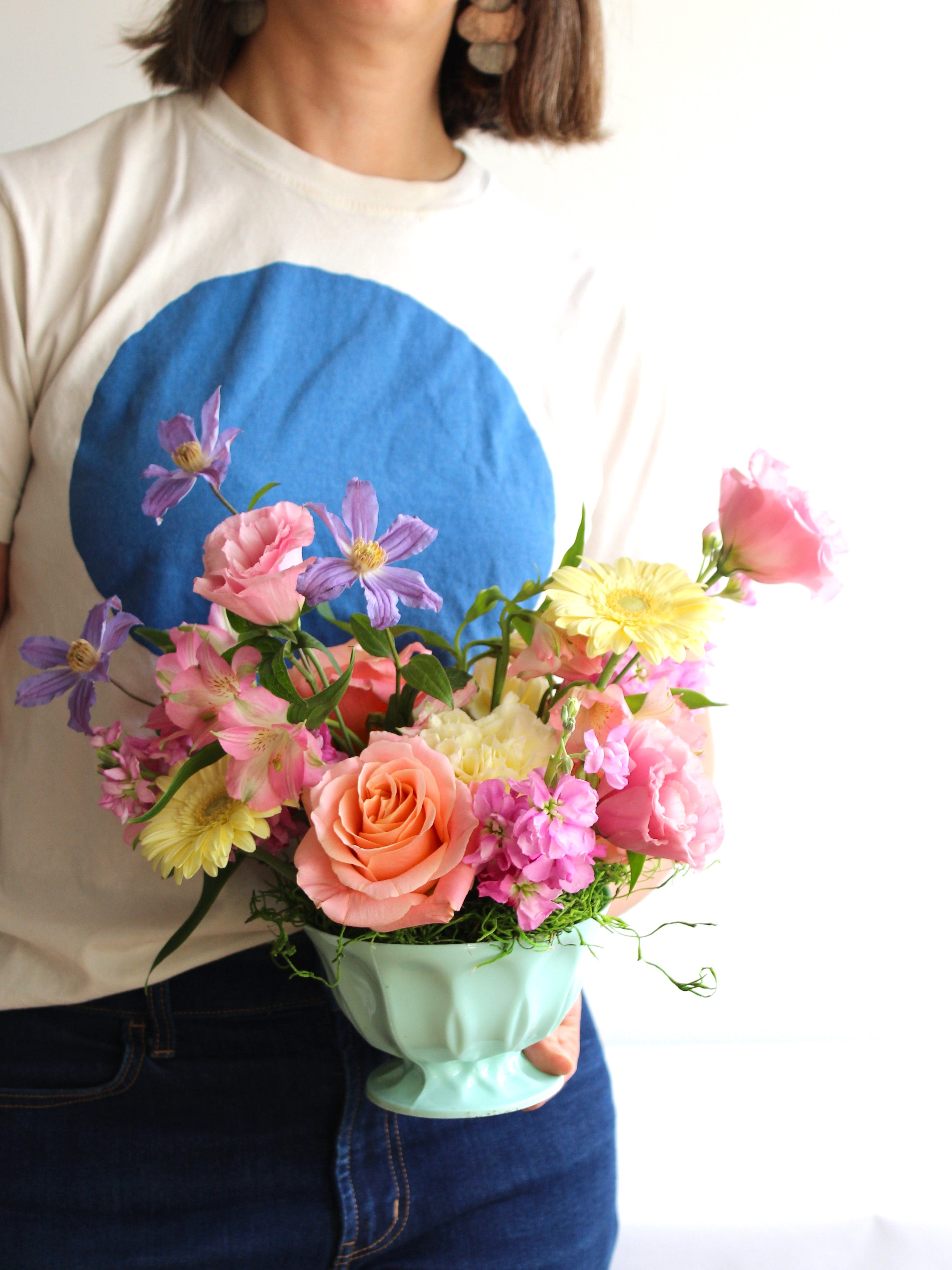 A woman wearing a t shirt with a blue circle on the front is holding an arrangement of pastel flowers.