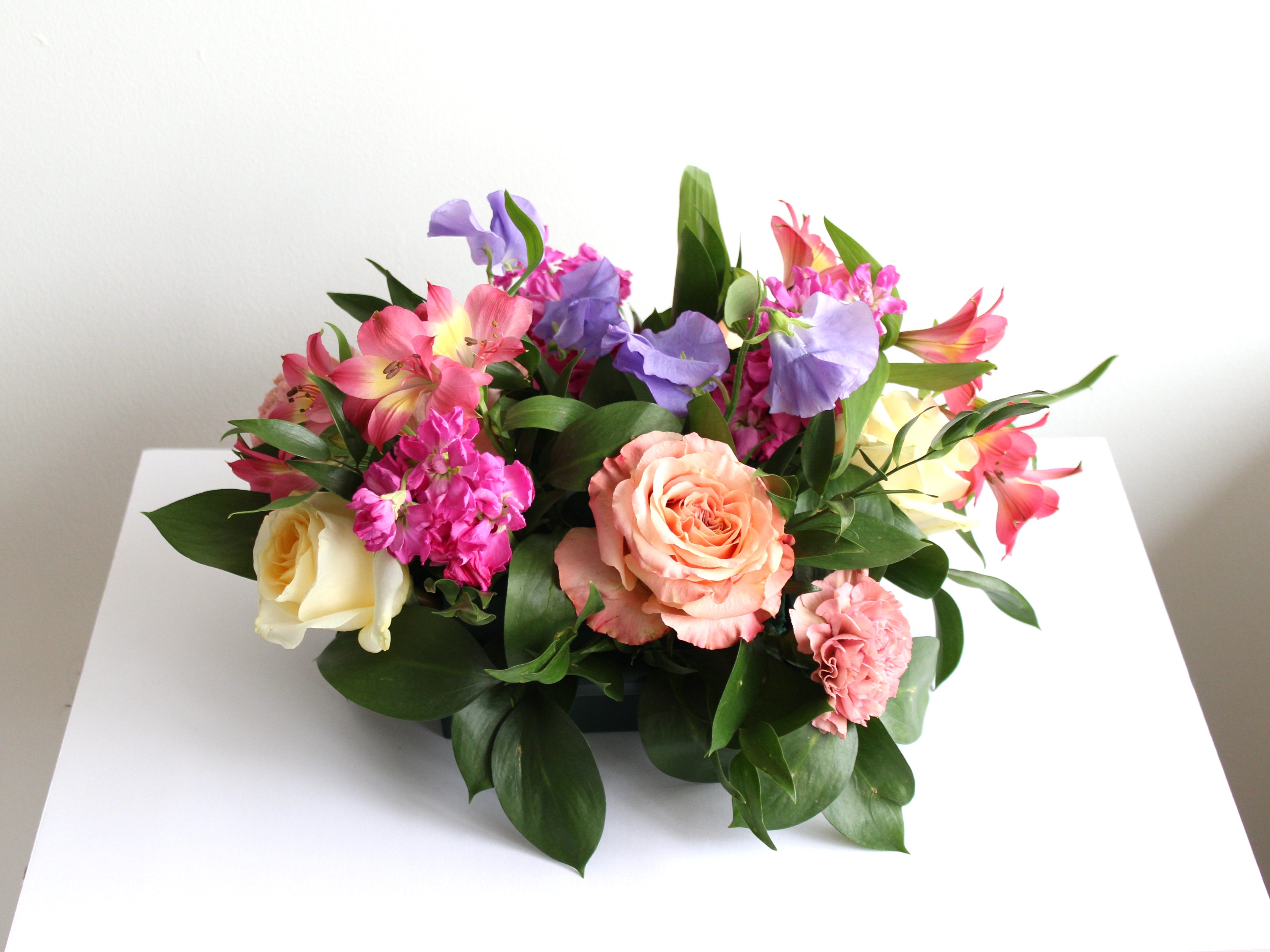 A low colourful arrangement of flowers in purple, pink, peach and soft yellow sitting on a  plain white tabletop.