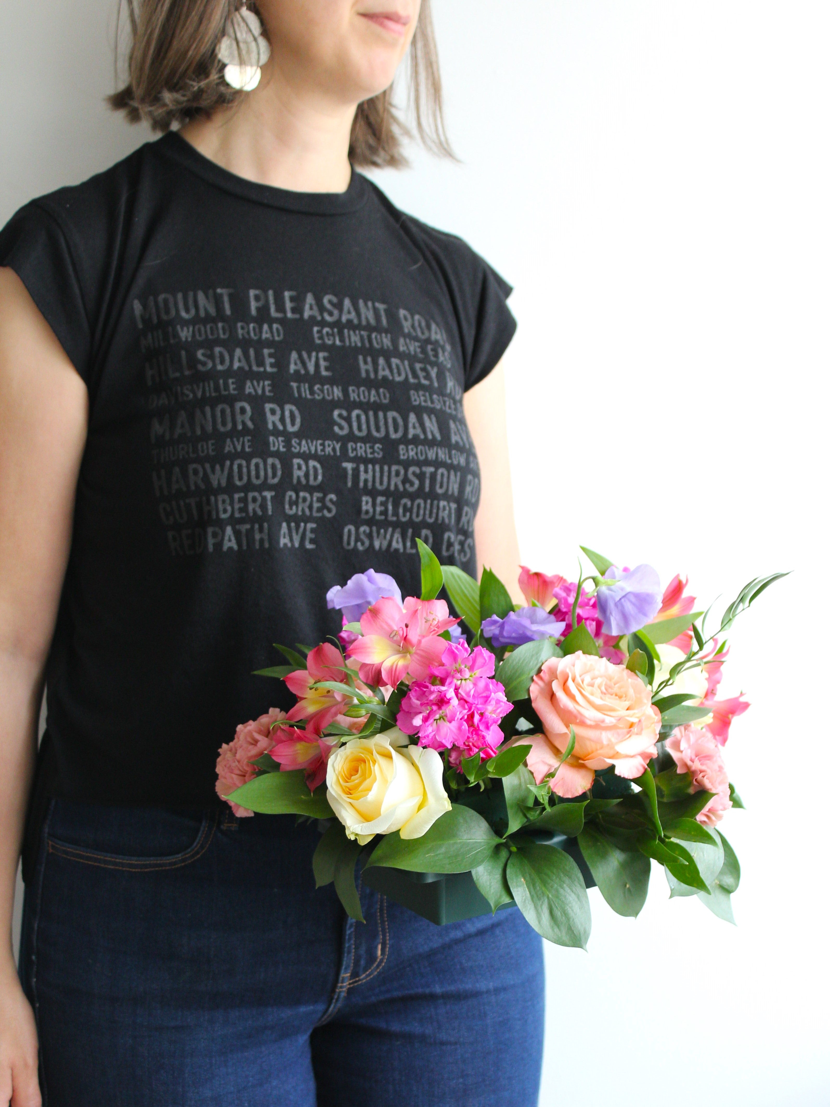 Toronto florist Periwinkle Flowers is holding a small low floral centerpiece, standing in front of a white wall.