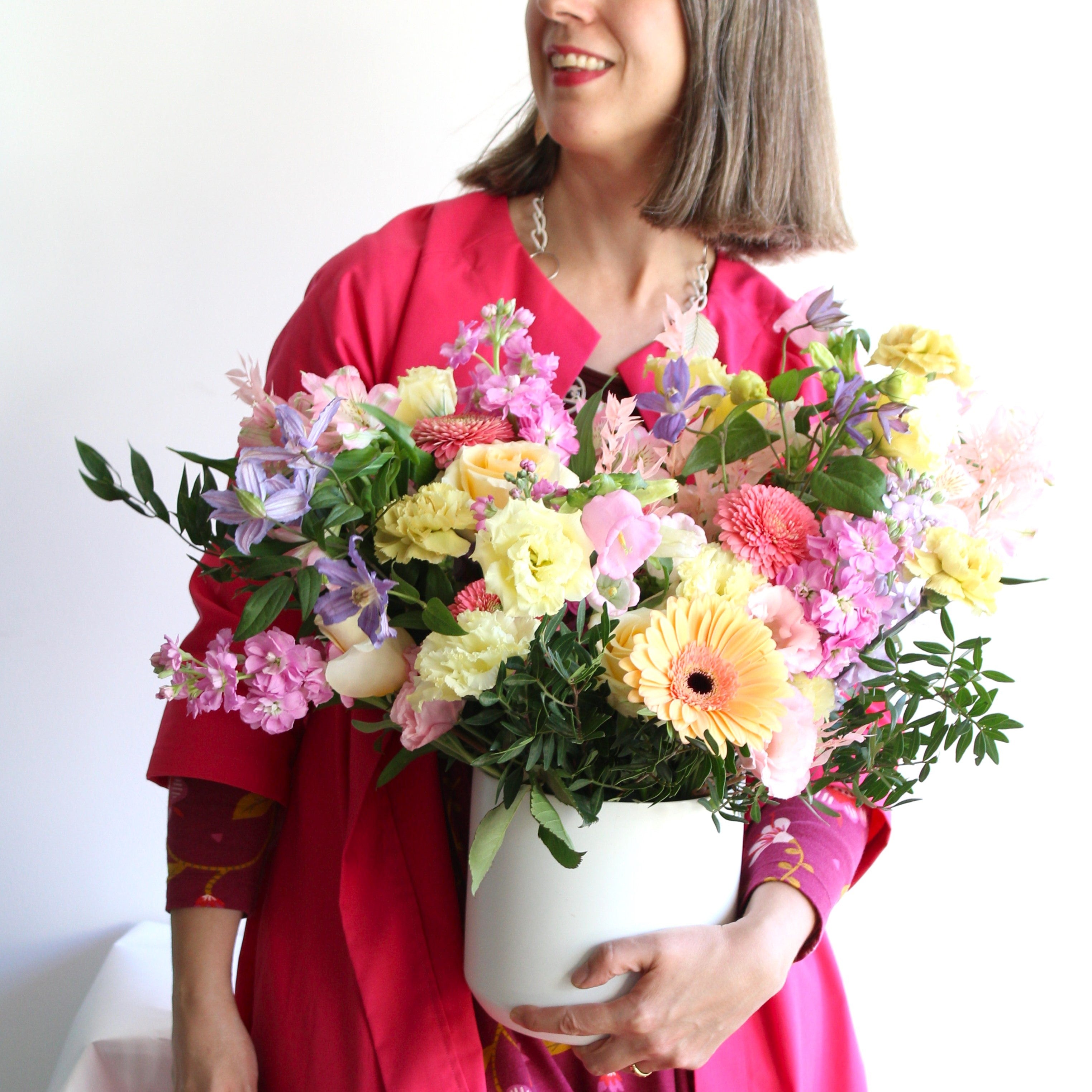 A woman is holding a white ceramic pot filled with flowers in pastel tones.