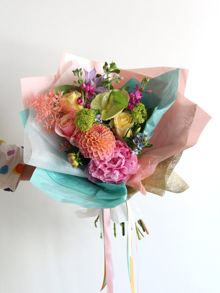 An arm is holding a mixed flower bouquet in pretty colours that is wrapped in decorative tissue paper.