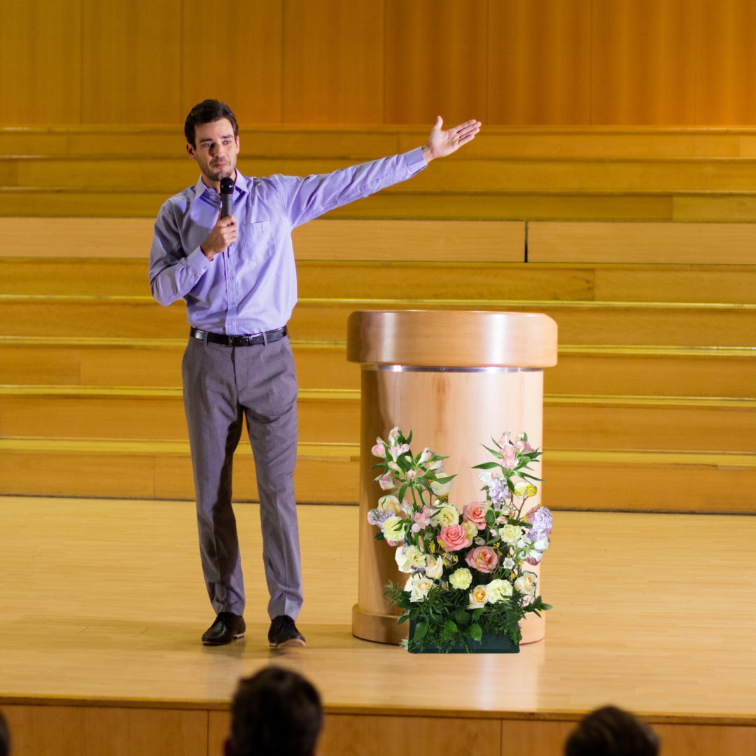 A man standing on a stage giving a speech, standing next to a lectern with a flower arrangement set in front.